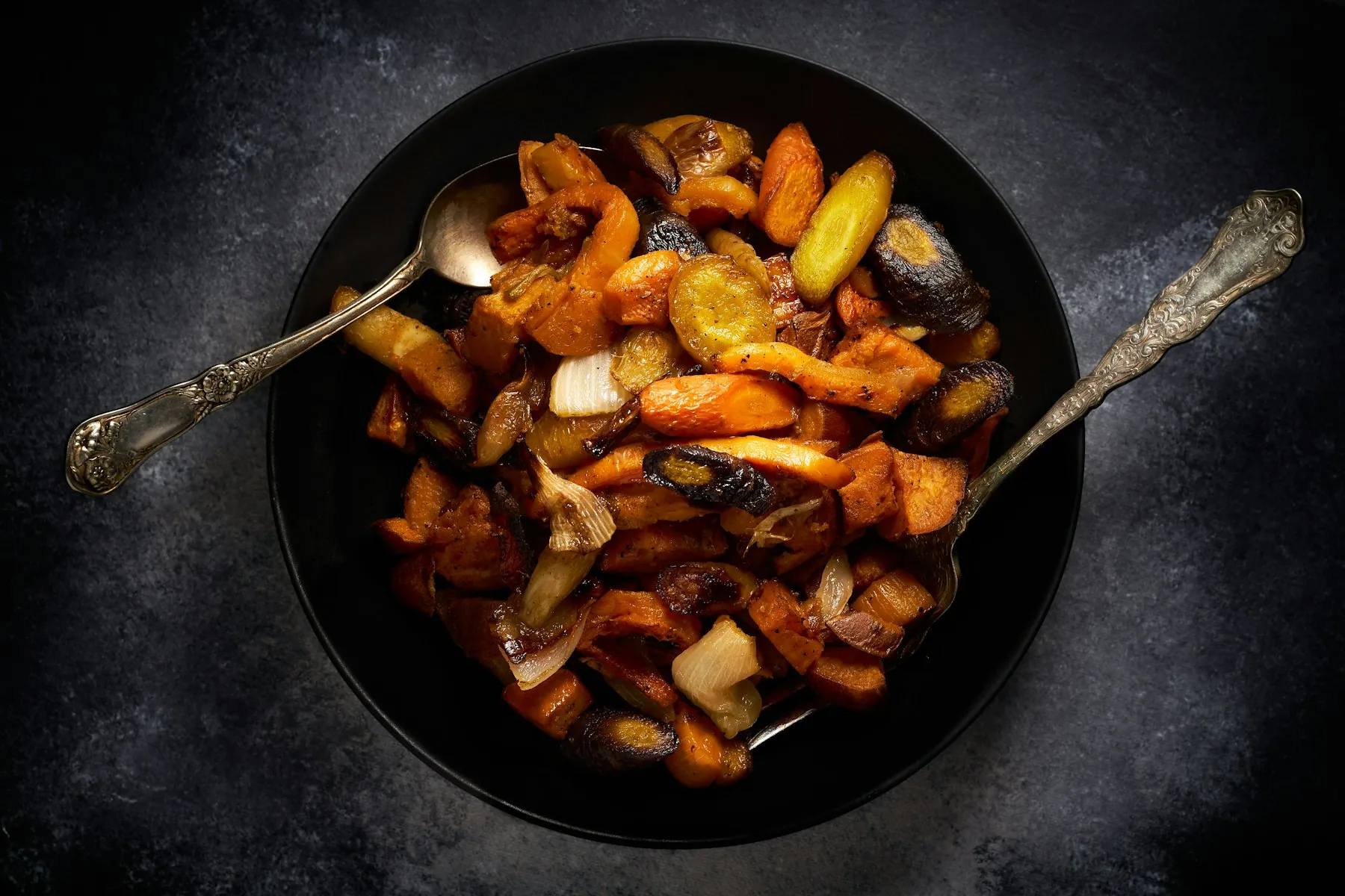 A black bowl of roasted heirloom carrots with two vintage spoons