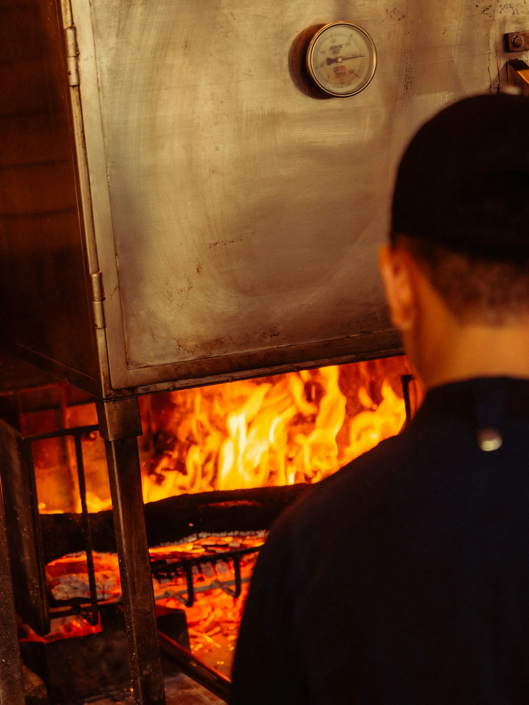 A cook in a black cap watching a wood fire inside an open oven