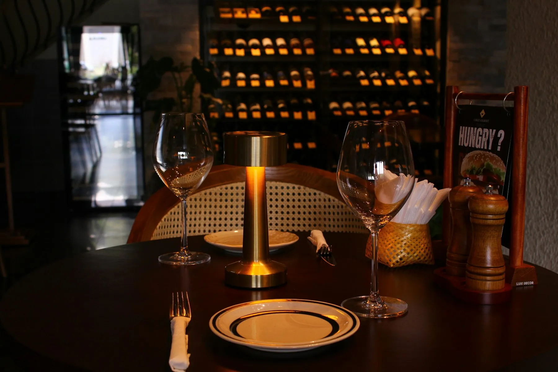 A table set with wine glasses, a lamp and a bottle rack behind